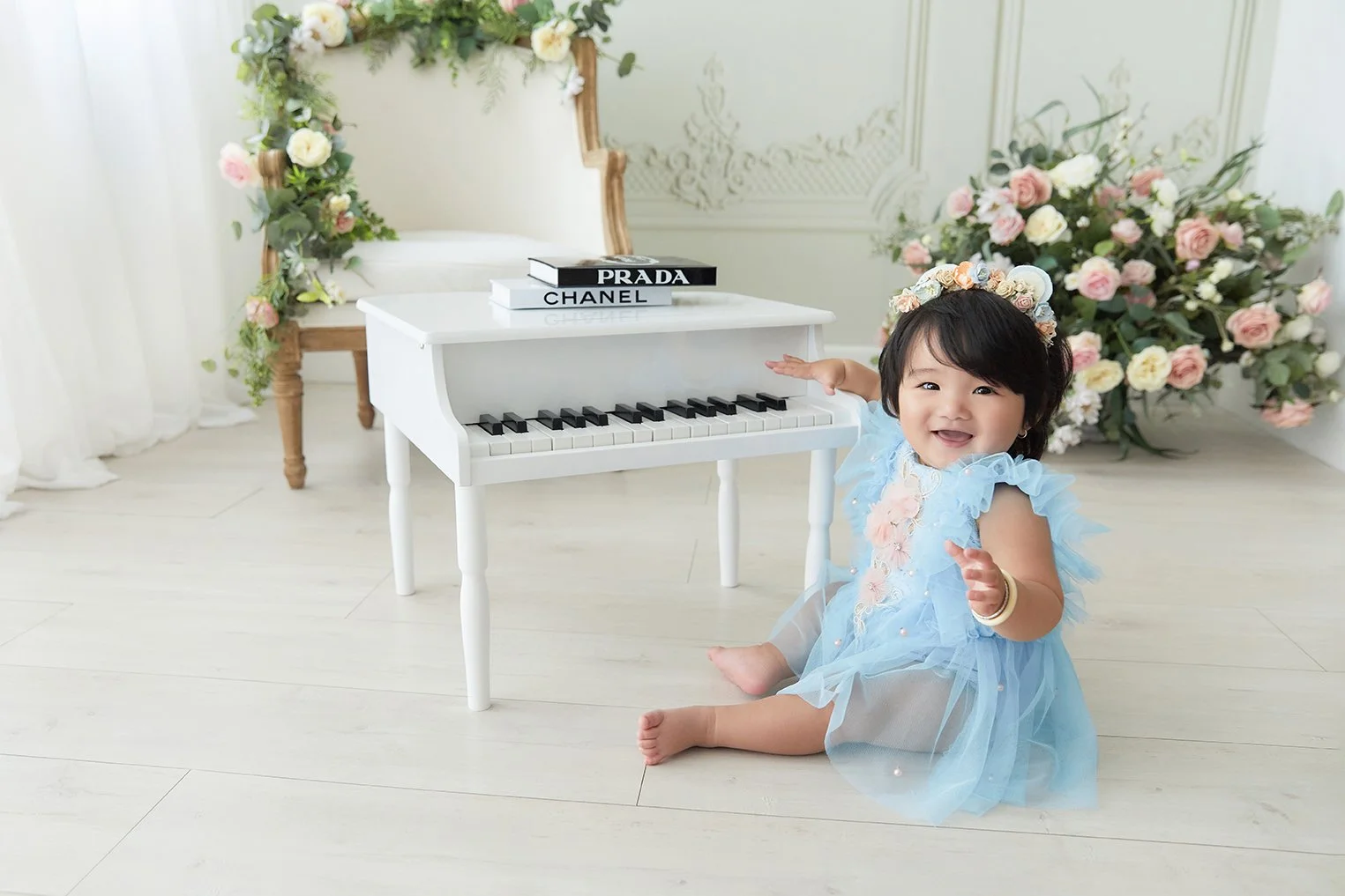 A young girl sitting on a light-colored wooden floor with a joyful expression, wearing a blue dress and a flower crown, next to a small white toy piano with books on top, surrounded by floral arrangements and white curtains.