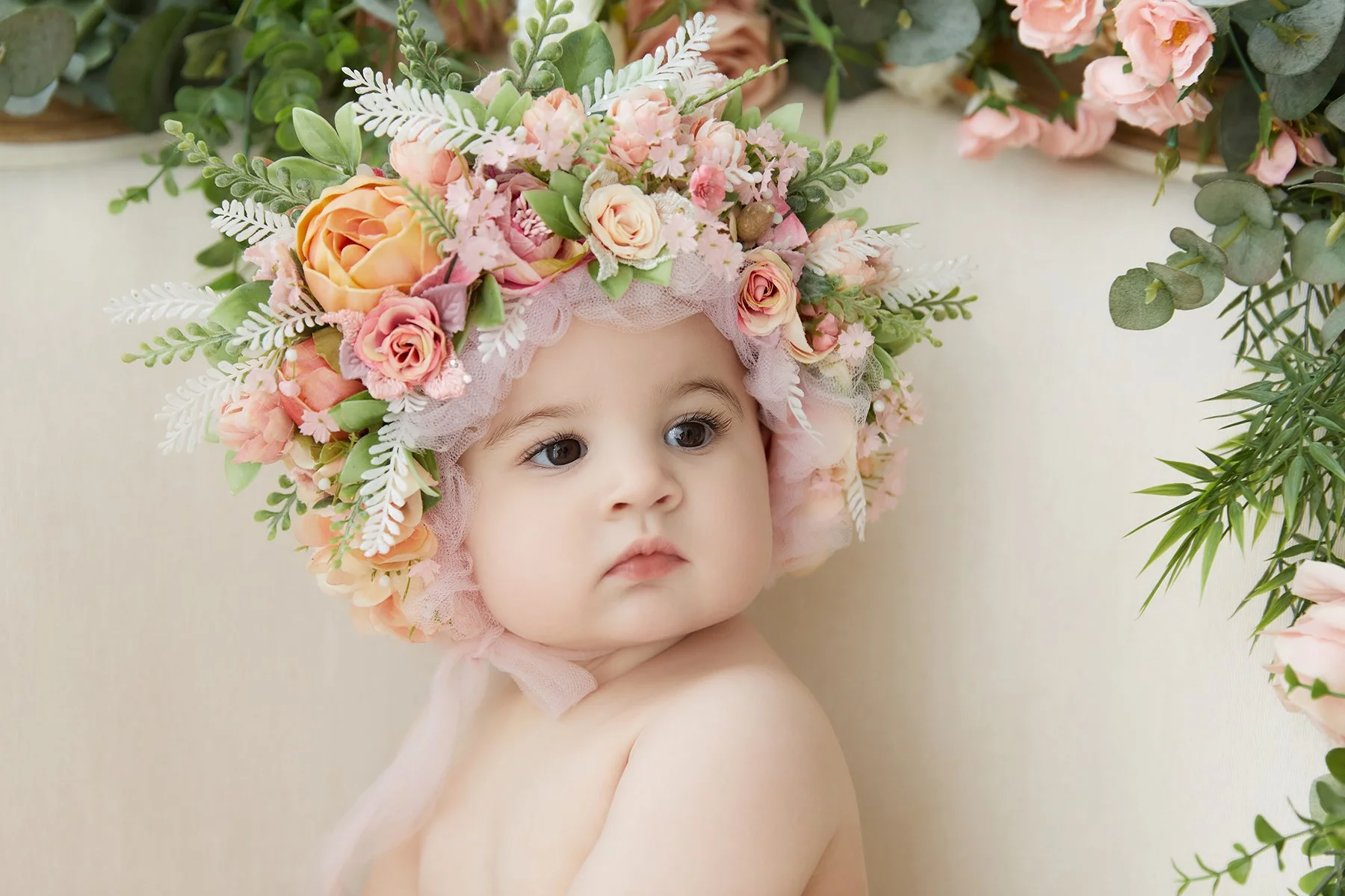A baby with light skin and dark big eyes, wearing a floral bonnet made of pink, peach, and white flowers, surrounded by greenery. The baby is sitting against a light neutral background.