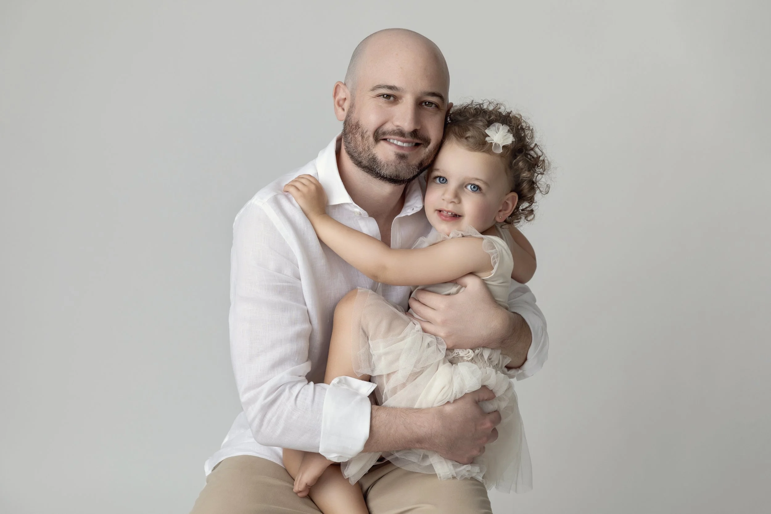 A smiling man with a beard holding a young girl with curly hair and a white bow, both in cream-colored dresses, against a plain light gray background.
