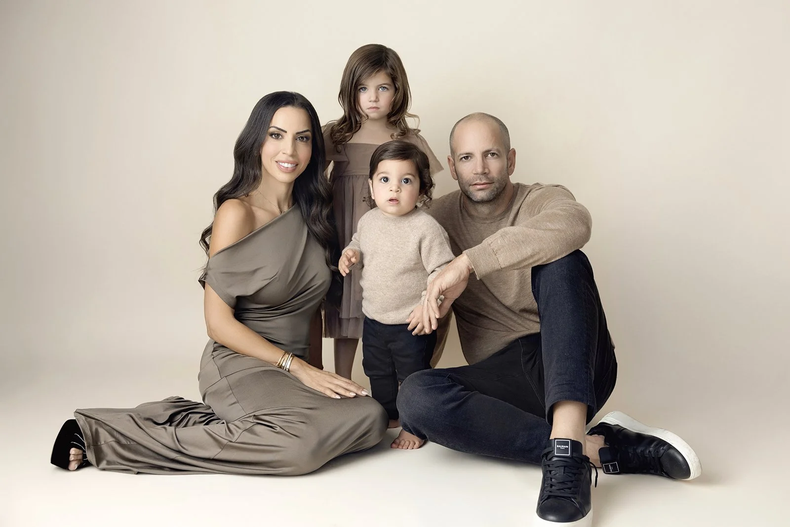 A family of four posing together in a photo studio with a plain background. The mother is kneeling on the left, wearing a taupe dress, with long dark hair. The father is sitting on the right, dressed in a beige sweater and black pants, with a shaved 