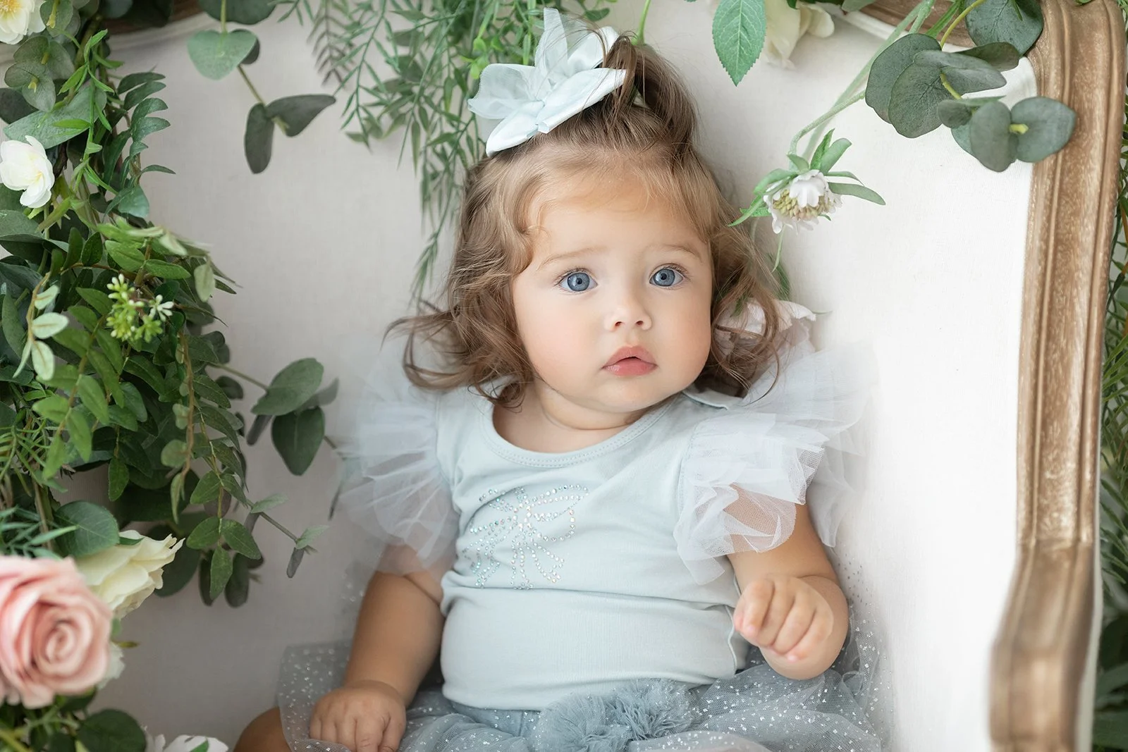 Young girl with curly hair, blue eyes, wearing a white top with tulle sleeves, sitting among green and white plants and flowers.