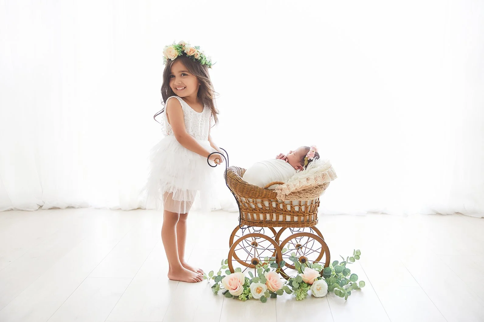 A young girl in a white dress and floral crown smiling while standing beside a vintage doll stroller with a sleeping baby girl inside, accessorized with a floral headband, surrounded by a flower arrangement on a white wooden floor.