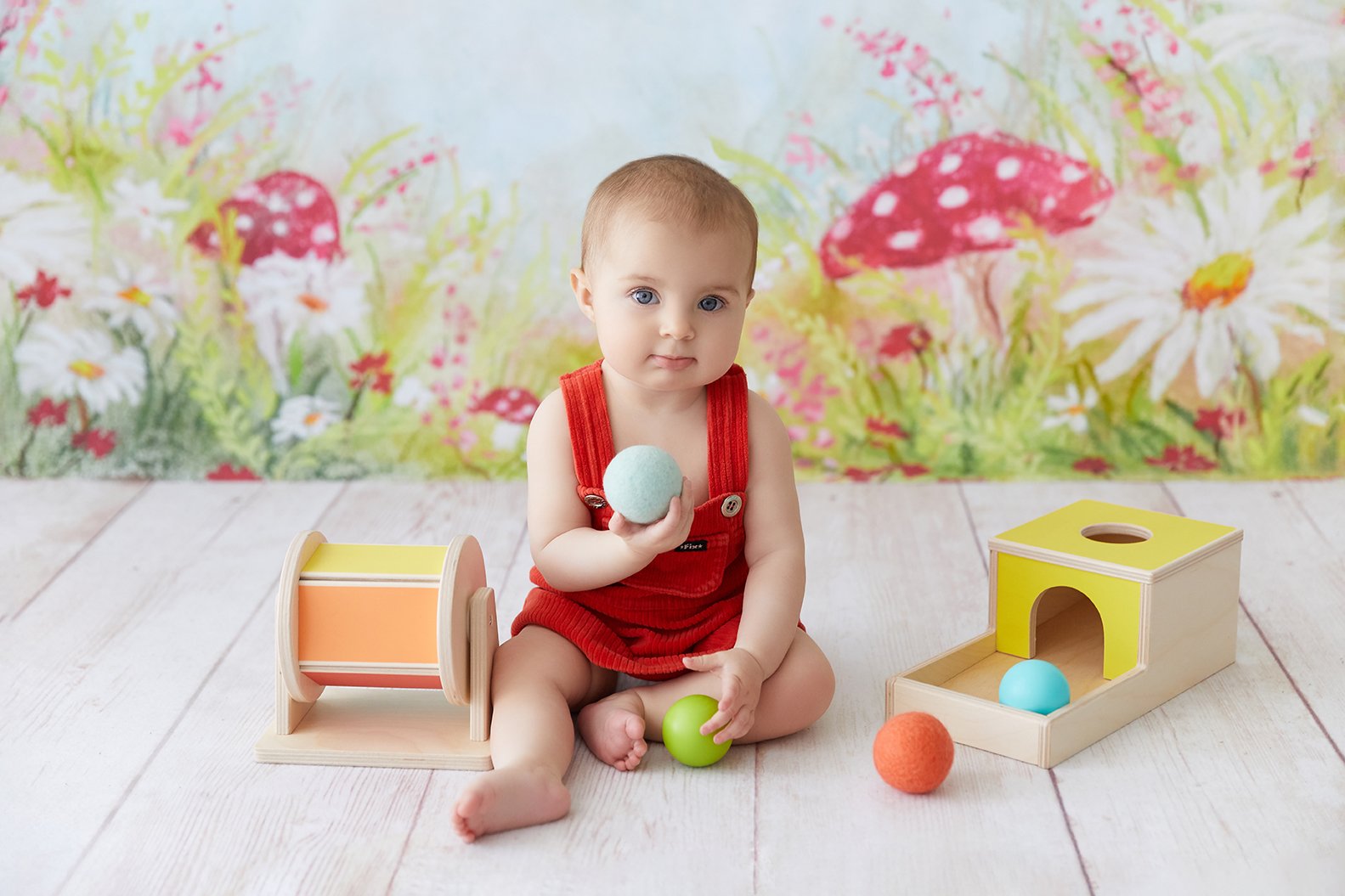 A young child with blue eyes and short hair, wearing red overalls, sitting on a white wooden floor, holding a light blue ball, surrounded by colorful wooden toys including a yellow and orange drum, a yellow and natural wood box with a blue ball insid