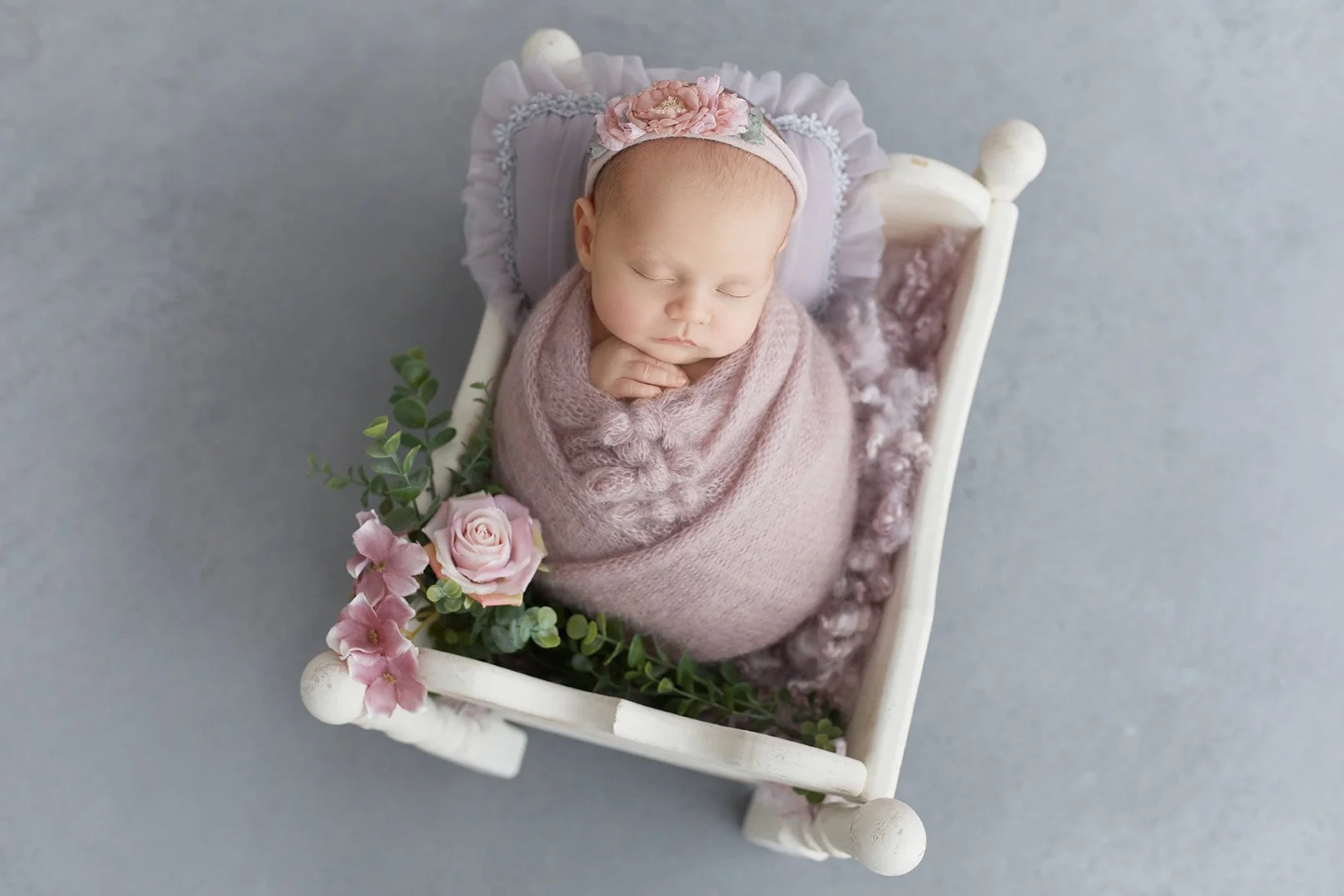 A sleeping infant wrapped in pink swaddling, lying in a small white wooden bed with floral decorations and pink roses.