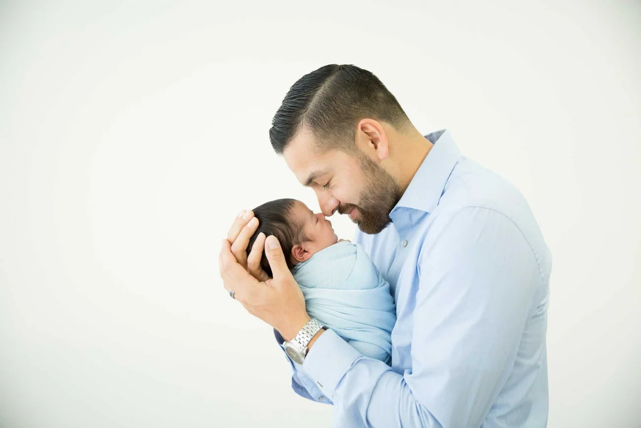 A man holding a newborn baby close to his face with foreheads touching, both smiling, against a plain white background.