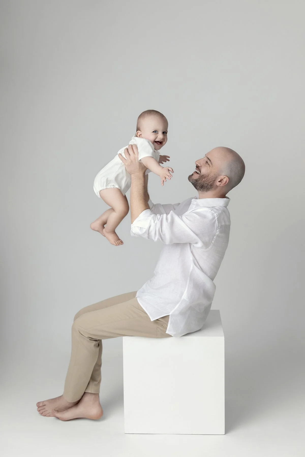 A man with light skin and a bald head sitting on a white cube, holding a smiling baby girl with light skin and light hair being lifted in the air.