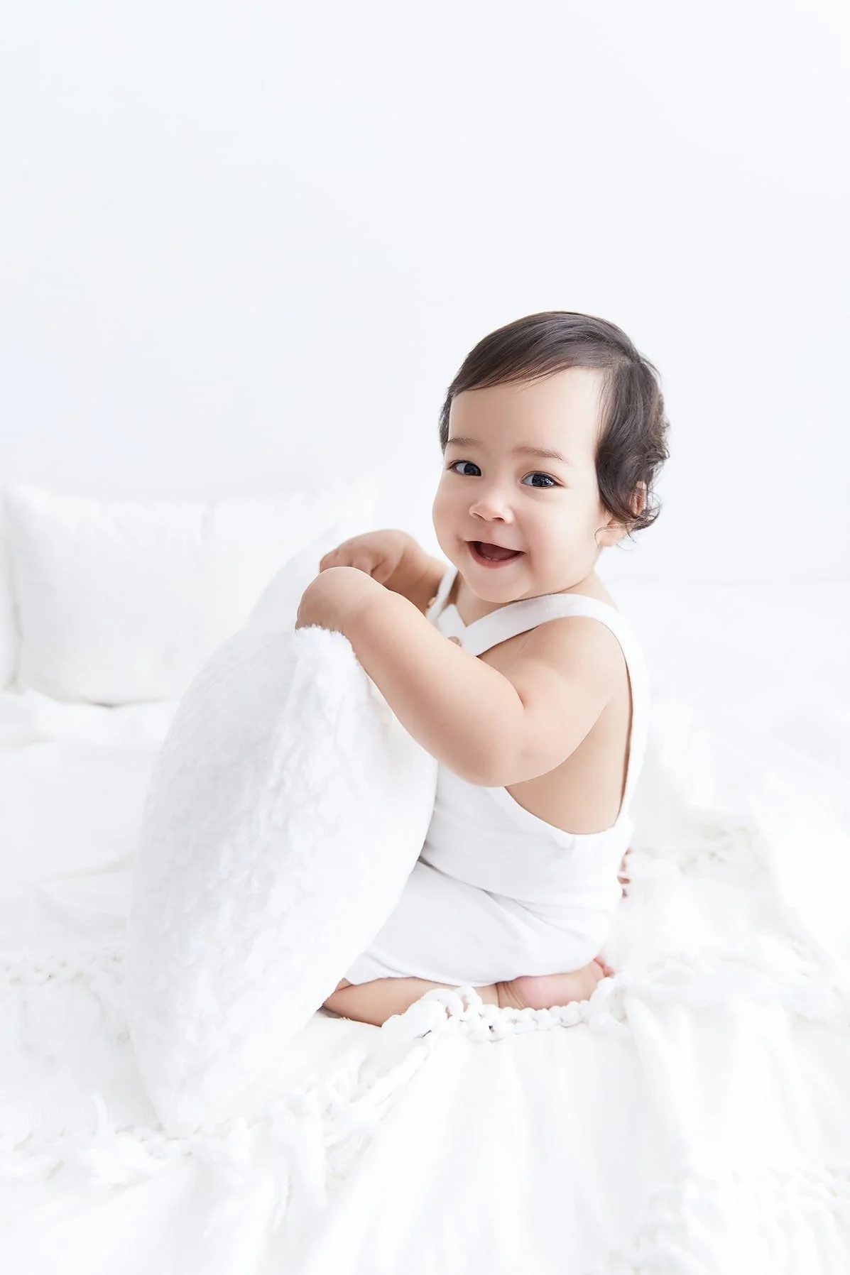 A smiling toddler with dark hair, wearing a white sleeveless outfit, sitting on a white bed while holding a white pillow in a bright, minimalist room.