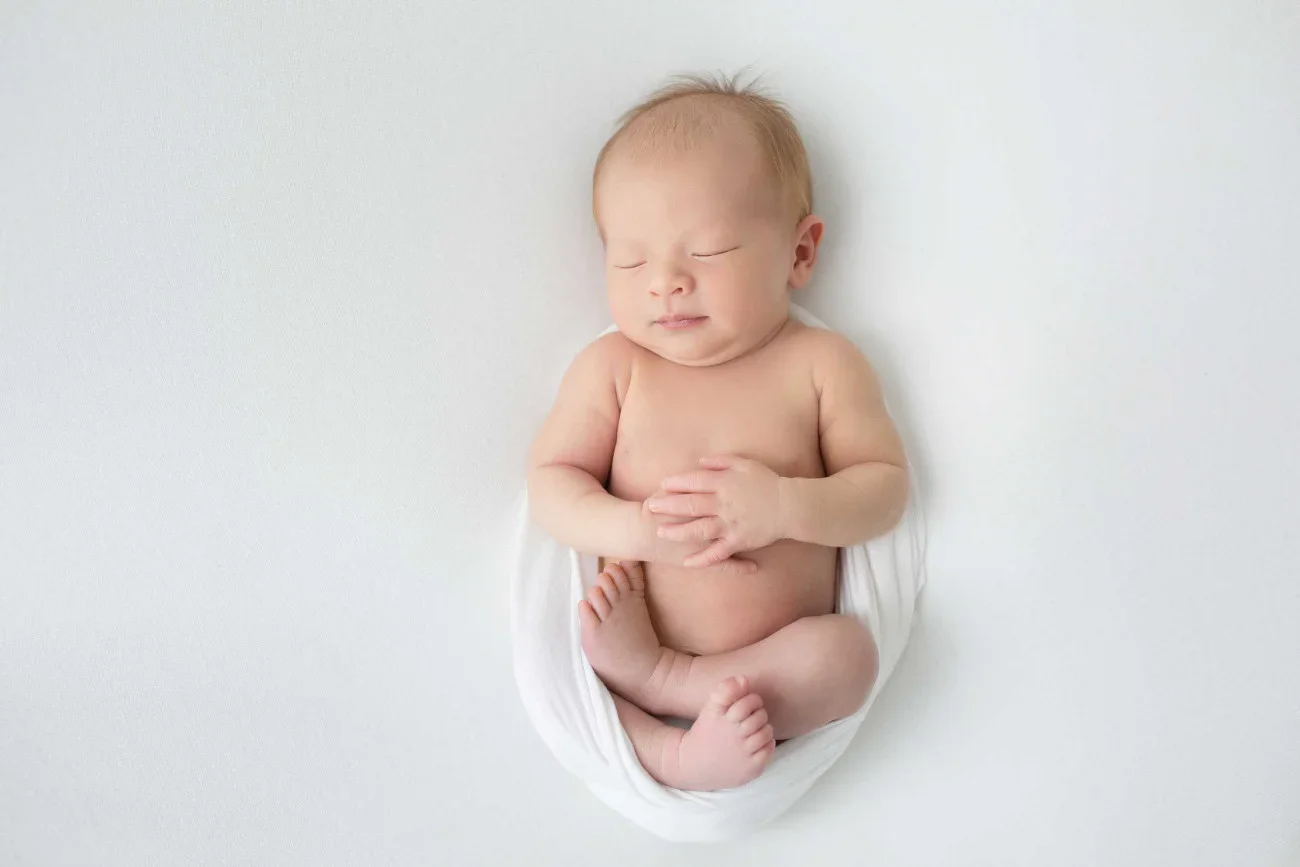A newborn baby with light skin and light hair, peacefully sleeping on a white surface, wrapped loosely in a white cloth.