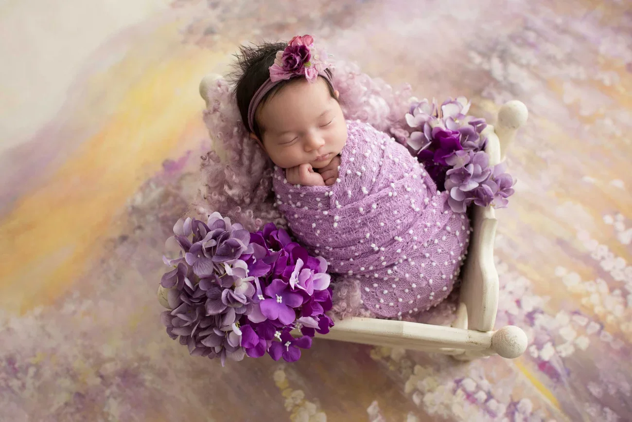 A sleeping baby wrapped in pink polka dot fabric, lying on a soft blanket in a small white bed decorated with purple and lavender flowers.