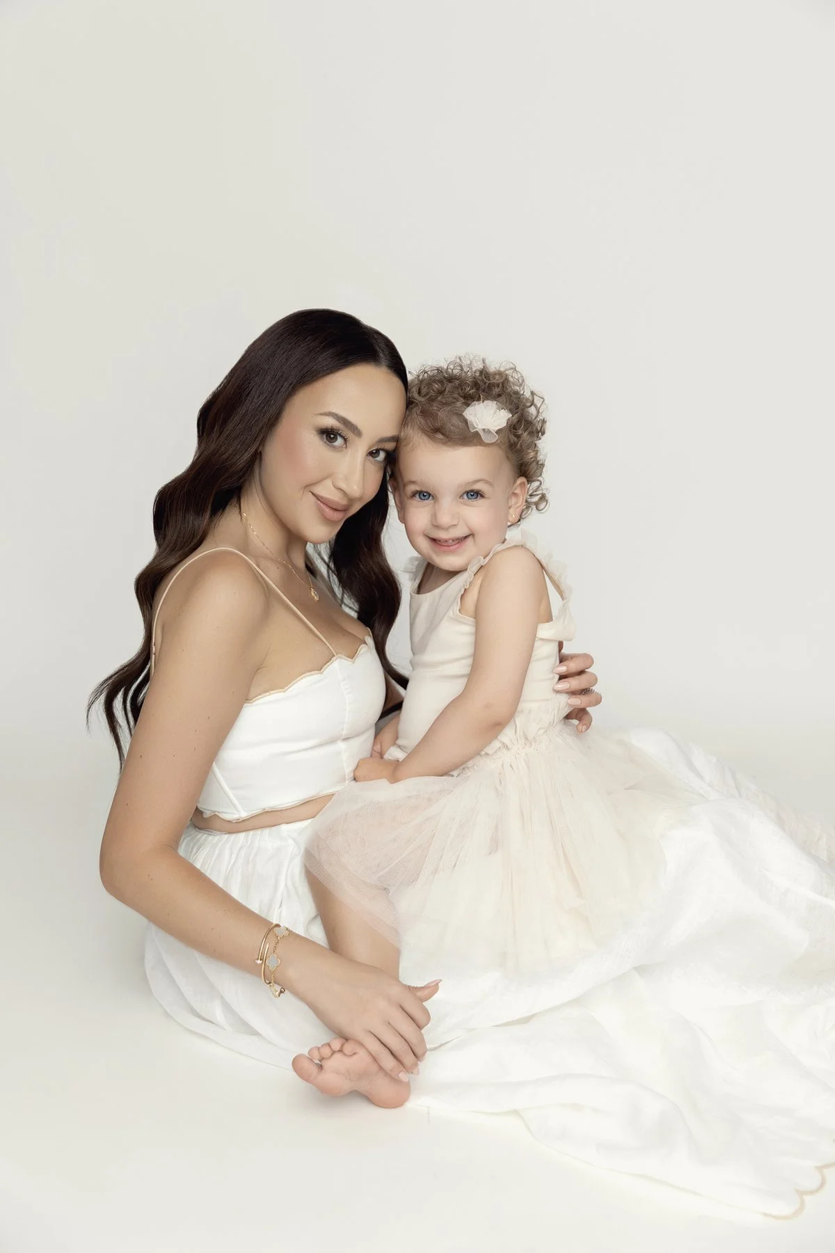 A woman and a young girl sitting together against a white background, dressed in white, smiling at the camera.