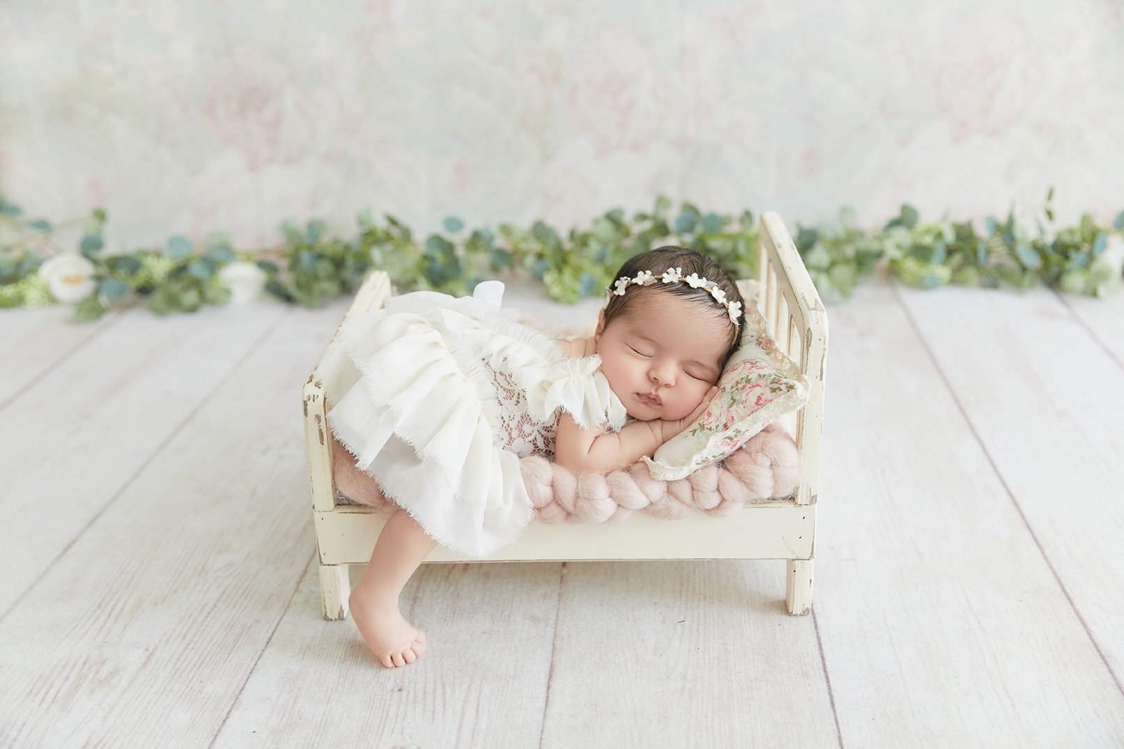 A sleeping baby girl lying on a small vintage bed with pink blankets, wearing a white lacy dress and a floral headband, on a wooden floor with green foliage in the background.