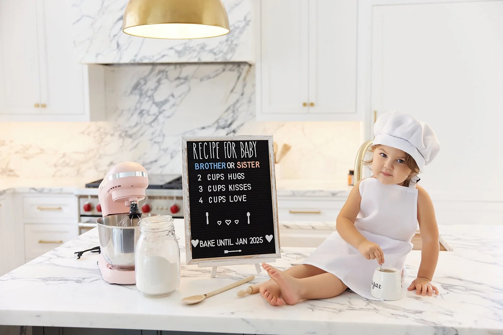 A young girl dressed as a chef sitting on a marble kitchen counter, stirring a cup, with a pink stand mixer, glass jar of flour, wooden spoon, and a blackboard with a baking recipe for a baby’s cake in the kitchen.