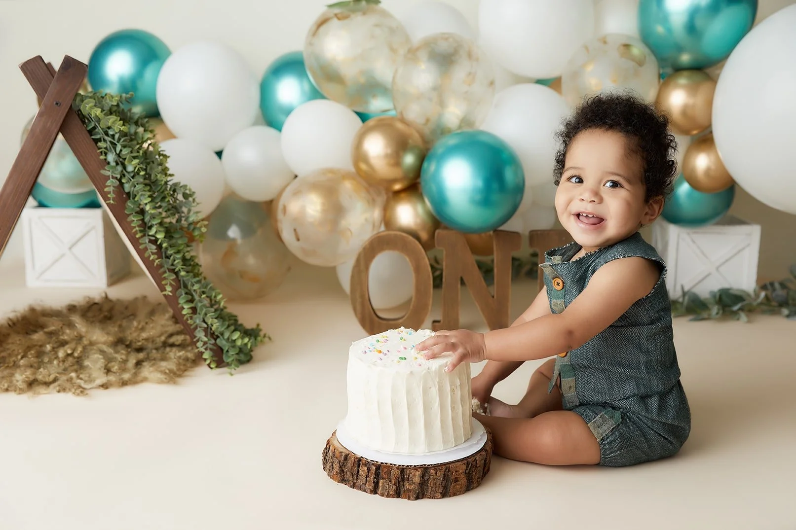 A smiling young child sitting on the floor near a white cake with sprinkles, celebrating a first birthday, with balloons and decorations in the background.