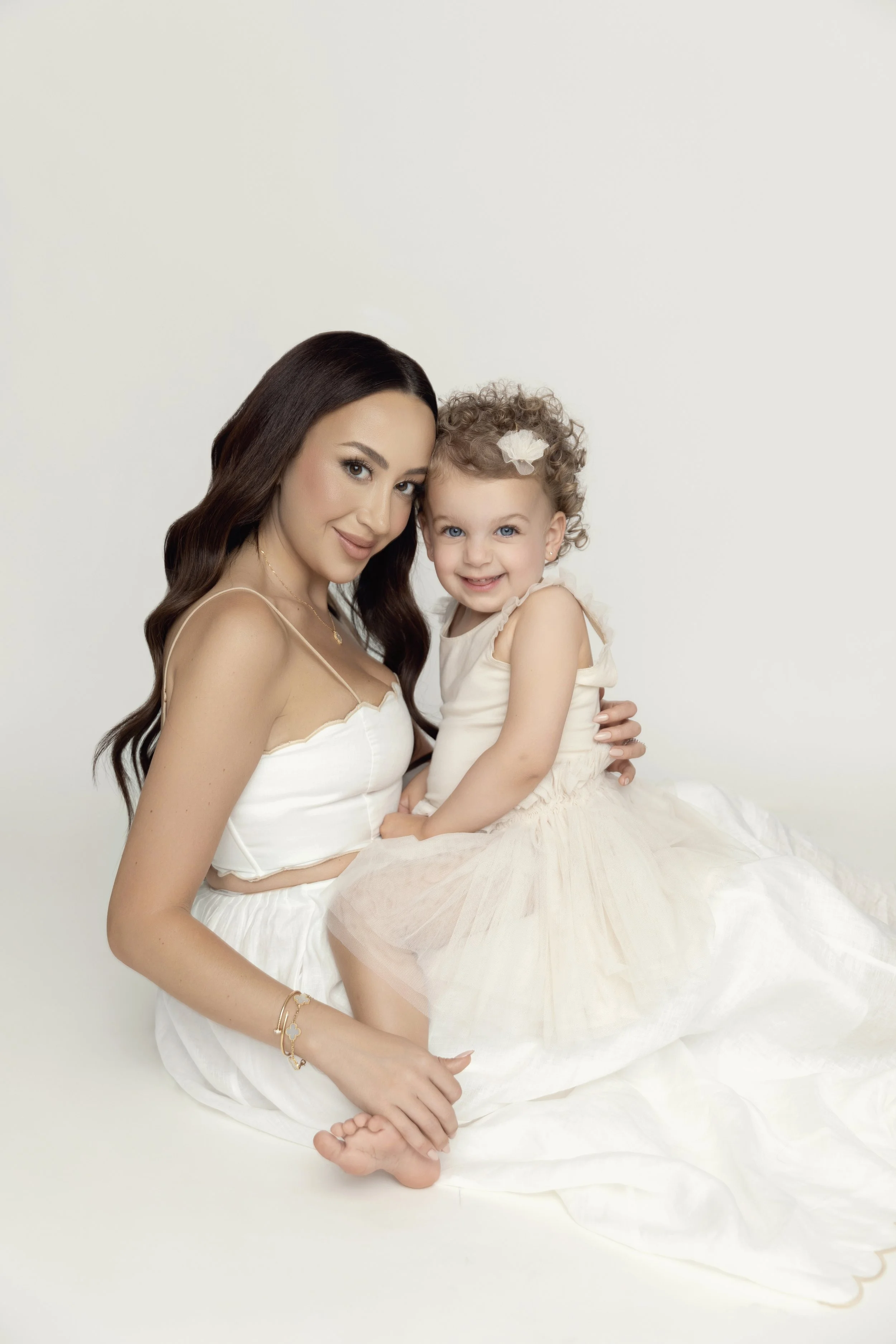 A woman and a young girl sitting together, both dressed in light-colored dresses, smiling at the camera against a plain white background.