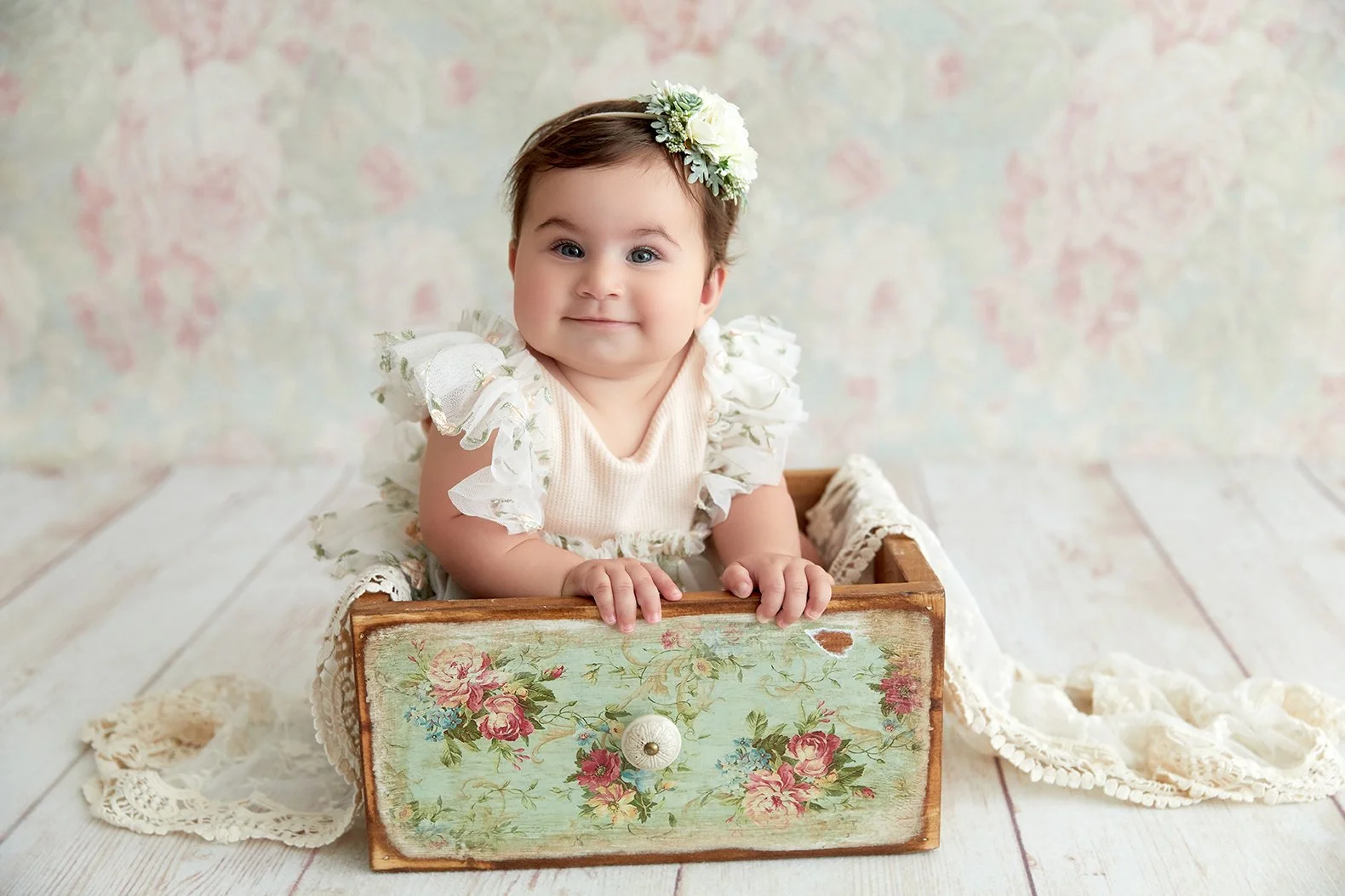 A smiling toddler girl with a flower headband, sitting in a wooden vintage box with floral design, on a wooden floor with a pastel-colored wall background.