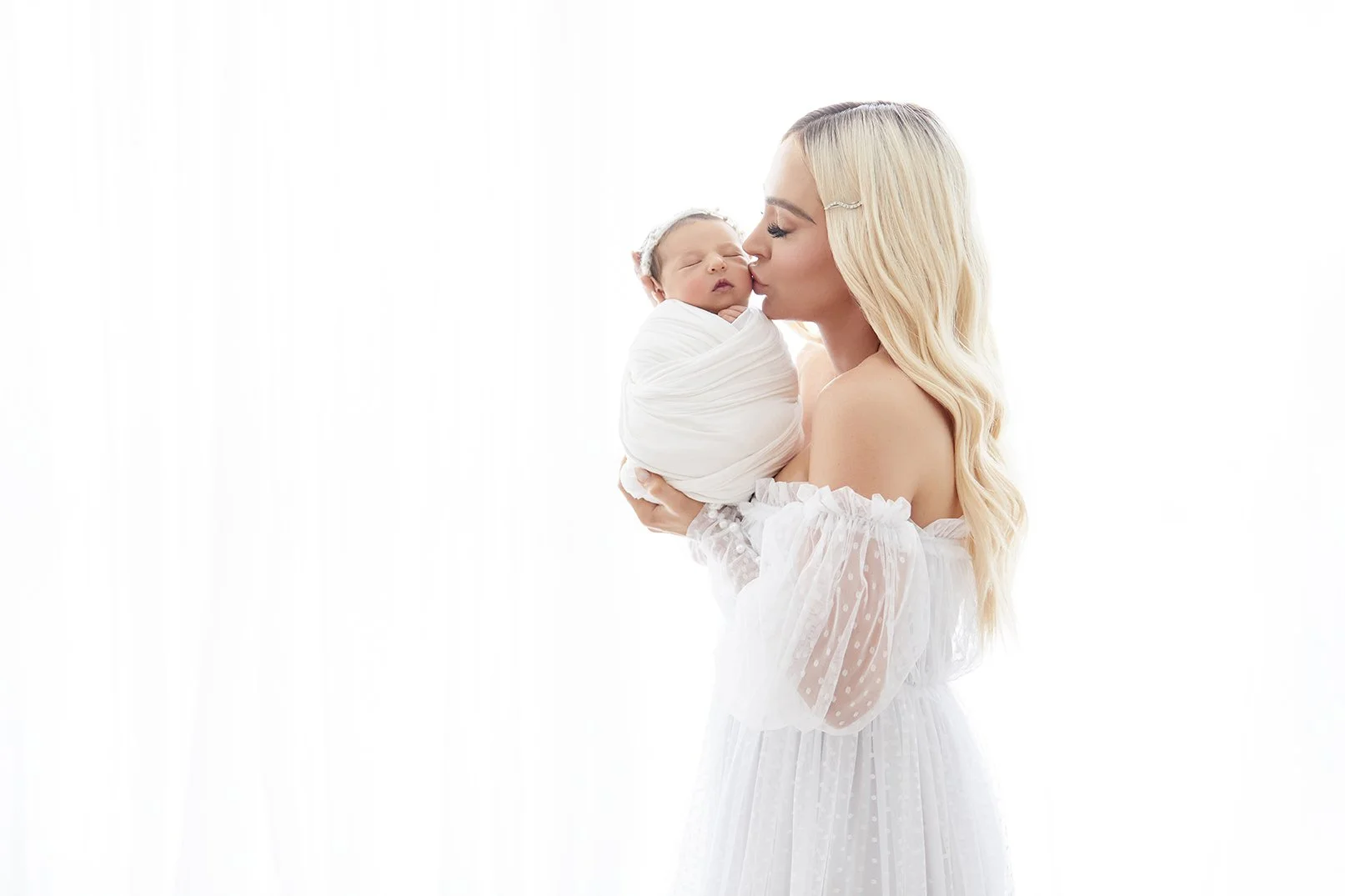 A woman holding a newborn baby in a white wrap, kissing the baby on the nose against a plain white background.