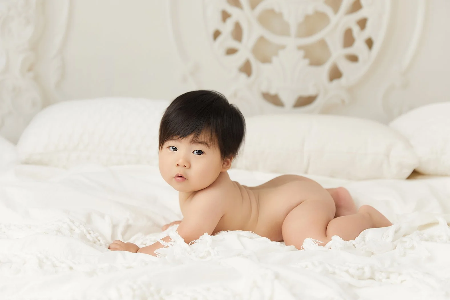 A young child with black hair lying on their stomach on a white bed, looking at the camera with a neutral expression.
