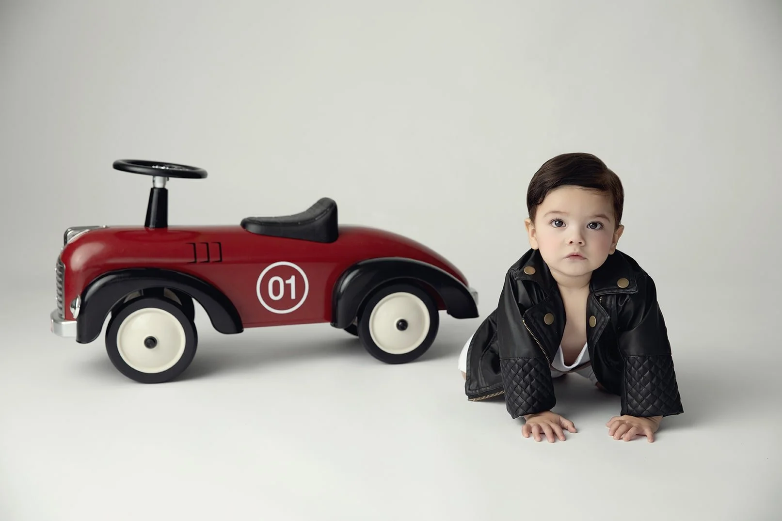 A young child crawling on the floor next to a red and black toy car, wearing a black leather jacket.