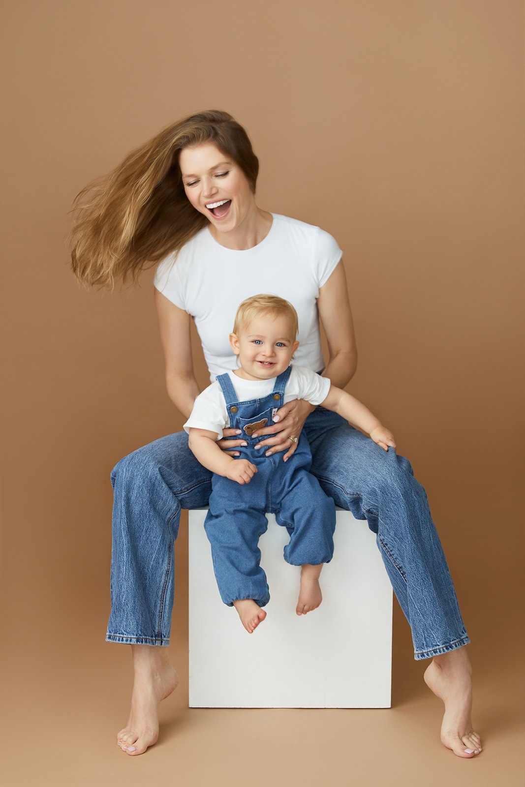 A woman and a young child sitting on a white cube against a brown background. The woman has long light brown hair, is smiling, and wearing a white t-shirt and blue jeans. The child, sitting on the woman's lap, has blonde hair, blue eyes, and is smili