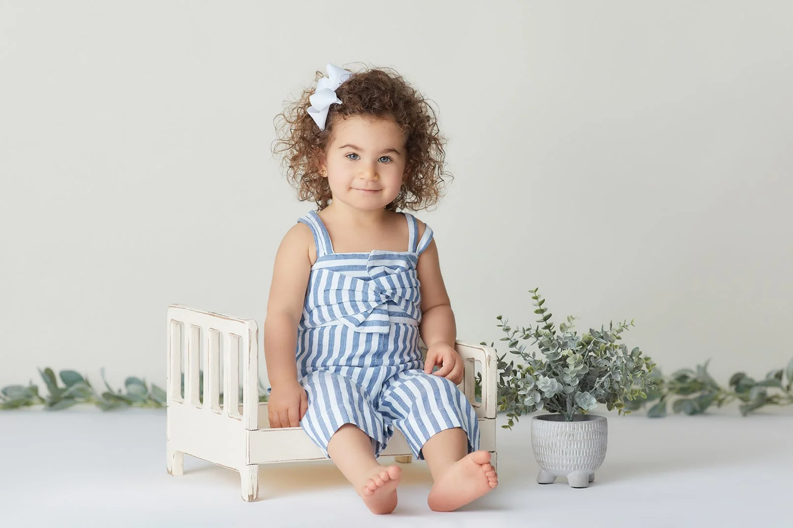A young girl with curly hair and a white bow, sitting on a small white wooden bed, wearing a blue and white striped romper. She is sitting barefoot next to a potted eucalyptus plant against a plain light background.