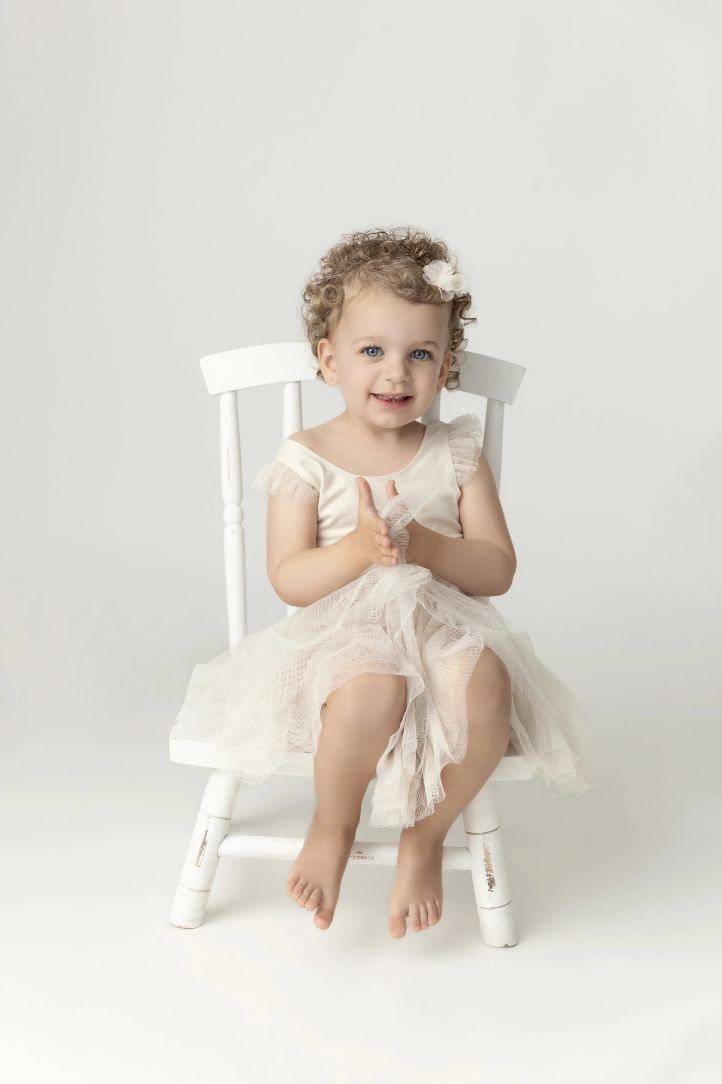 Young girl with curly hair sitting on a white wooden chair, wearing a cream-colored dress with tulle, clapping her hands, and smiling against a plain white background.