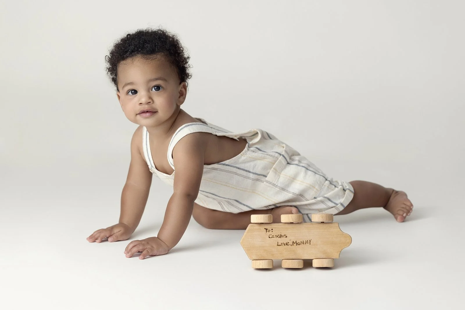 A young child with curly hair crawling on a white floor next to a wooden car-shaped block with a handwritten note reading 'To: Cassius Love, MoMMY.'