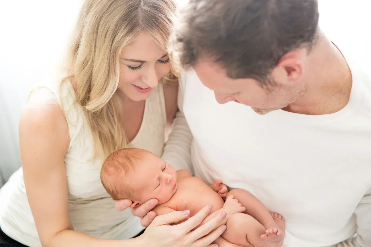 A family holding a newborn baby. The woman is smiling and looking at the baby while the man is looking at the baby, who is sleeping peacefully.