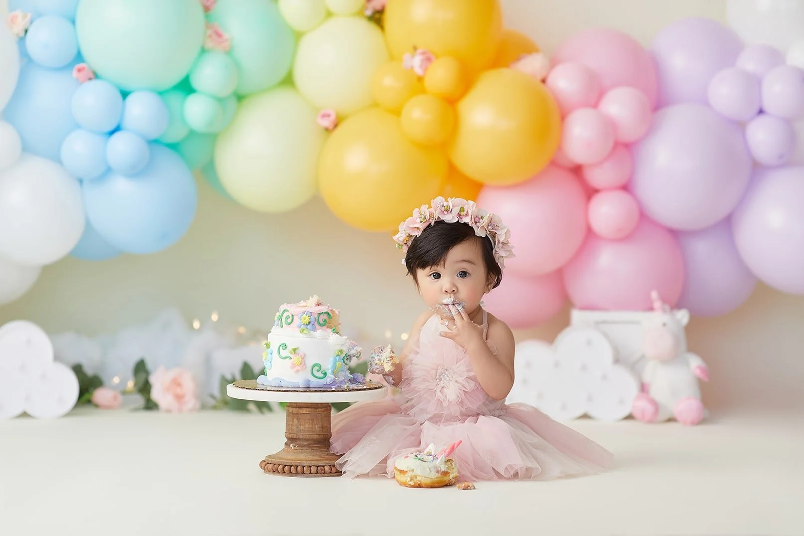 A young girl in a pink dress and flower crown sitting on the floor with a small cake in front of her, surrounded by pastel-colored balloons in the background, celebrating a birthday.