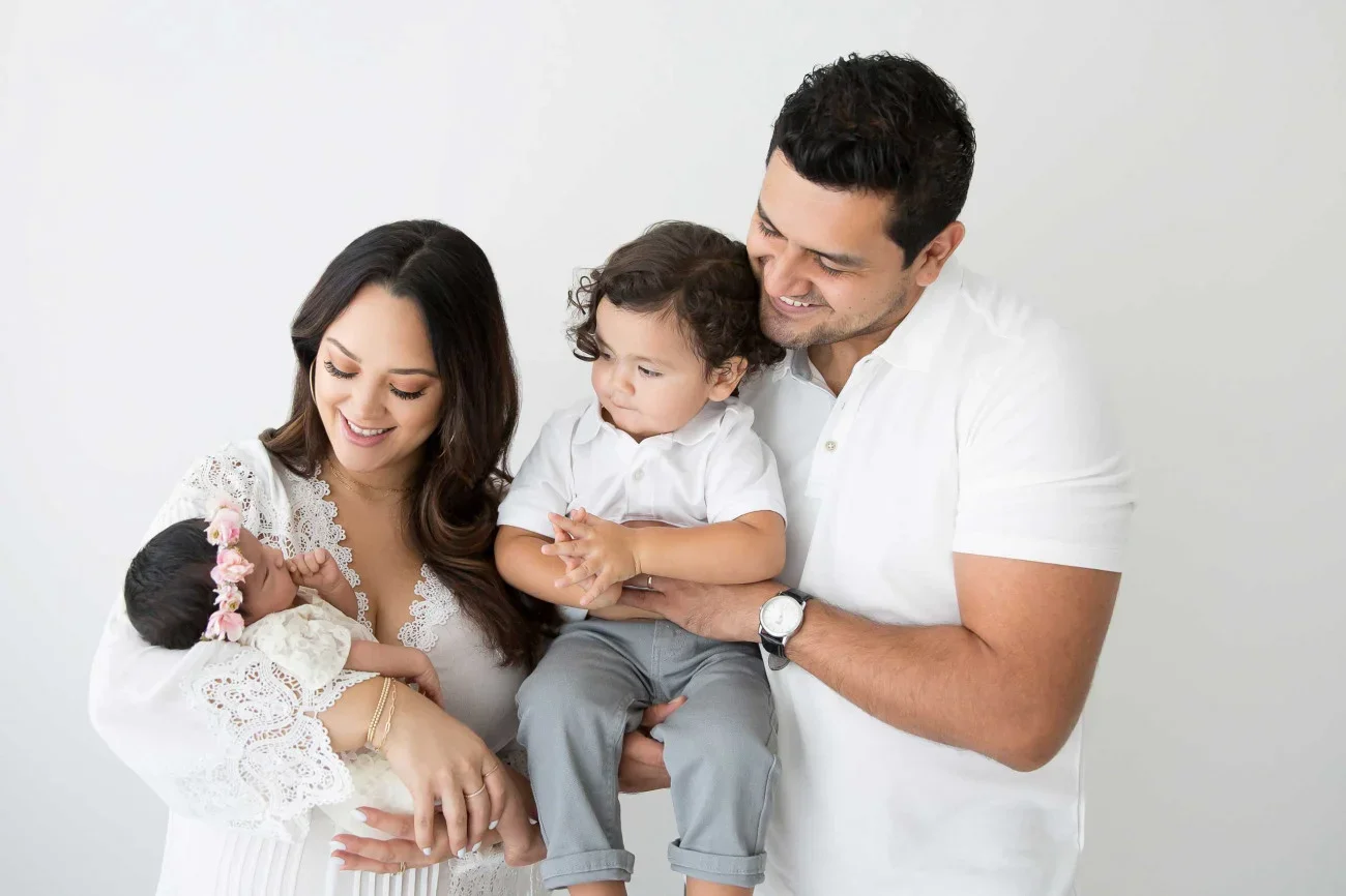 A family of four, including a mother, father, and two young children, standing together against a plain white background. The mother is holding a newborn girl dressed in white with a floral headband, while the older boy, standing between the parents,