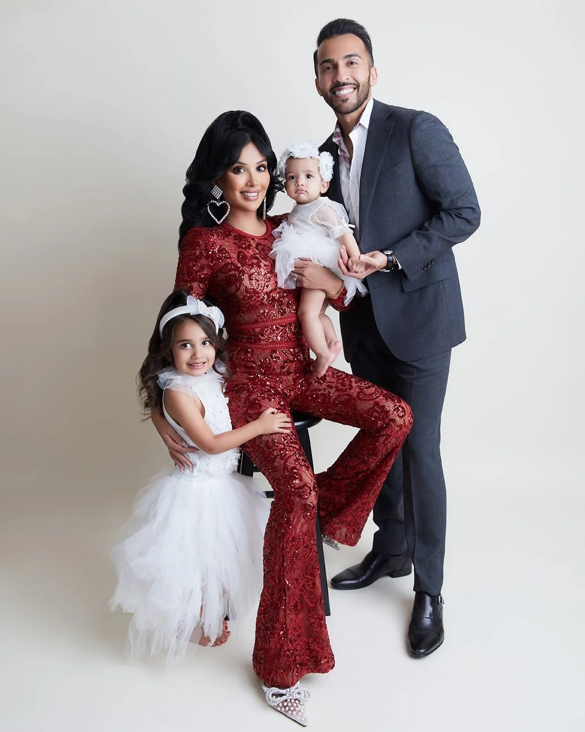 Family portrait of a woman in a red lace dress, man in a suit, and two young girls in white dresses, posing together.