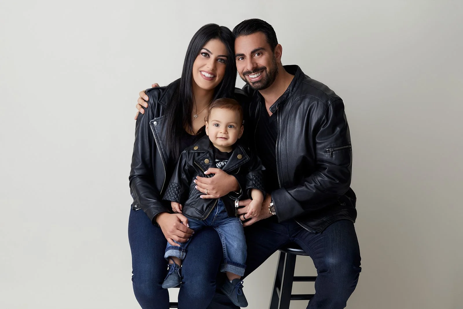 A family of three, a woman, a man, and a young boy, dressed in black leather jackets, sitting together against a plain background, smiling at the camera.