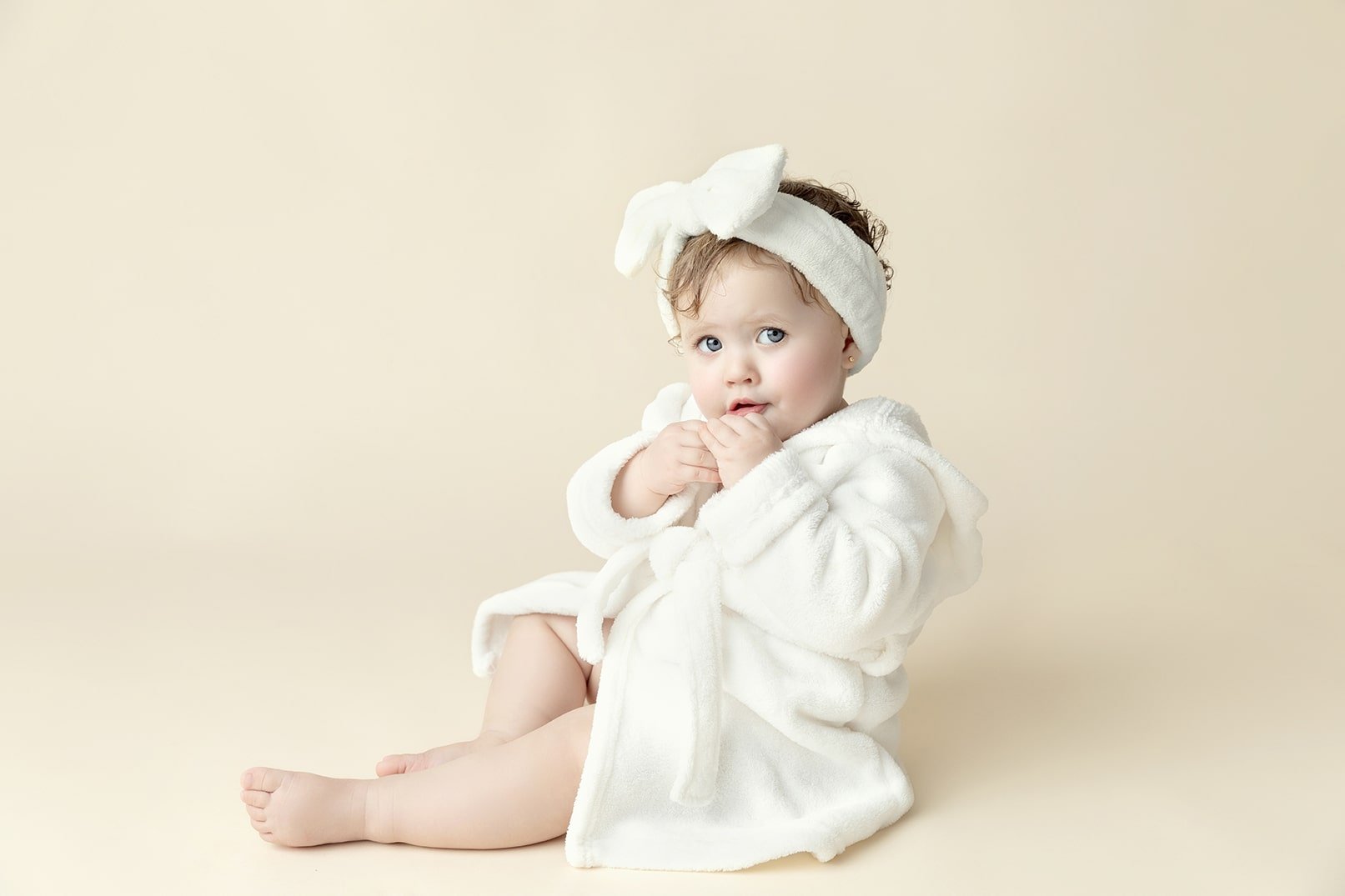 A toddler with blue eyes wearing a white bathrobe and a matching headband with a bow, sitting on a light beige background.