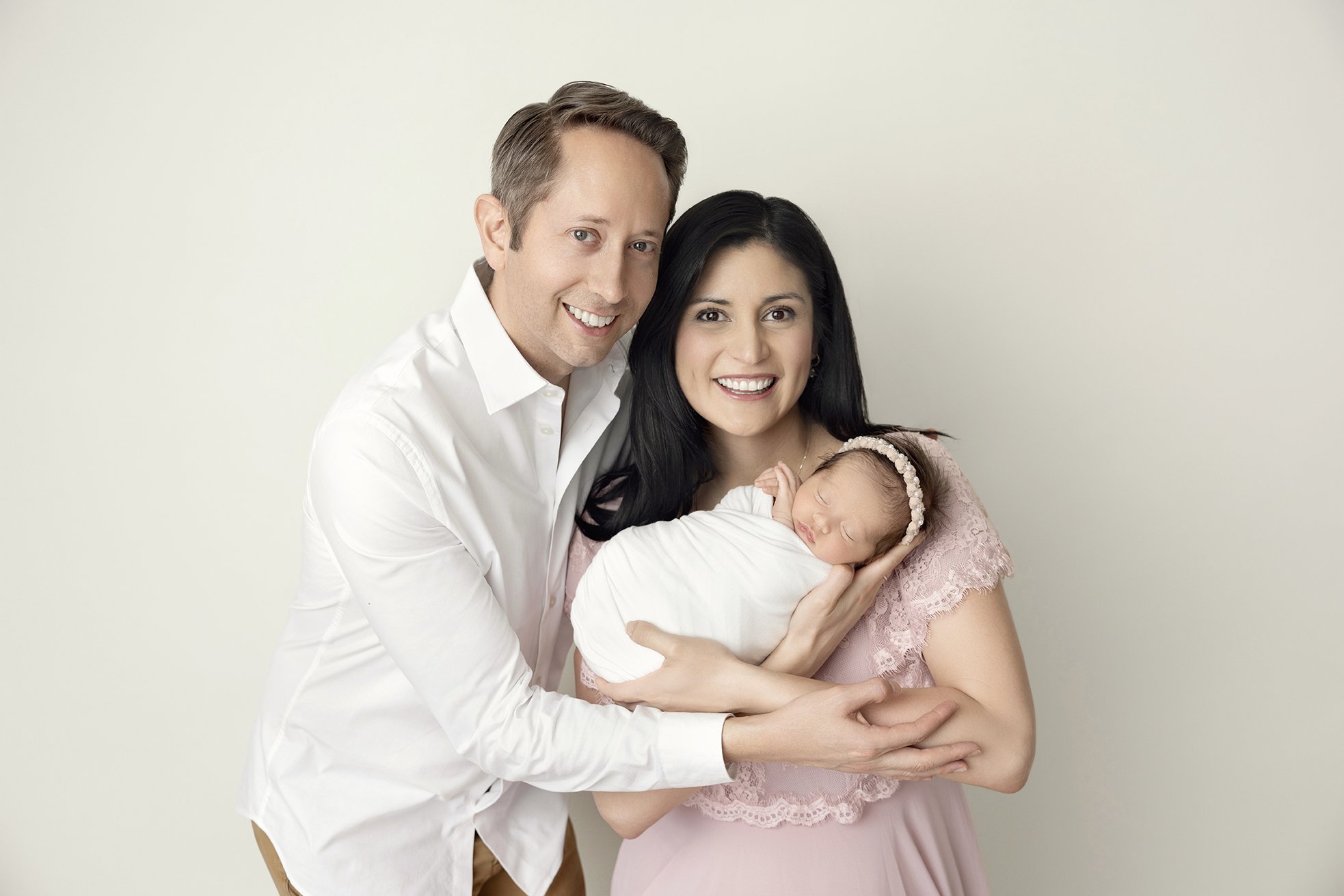 Happy family of three, parents holding their newborn baby, all smiling, in a studio with a plain background.