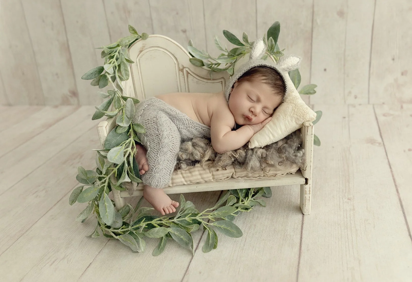 A sleeping newborn baby lying on a small vintage white bed with green leafy vine decorations, wearing a knit hat with bunny ears and cozy knitted pants, resting on a small pillow on a wooden floor background.