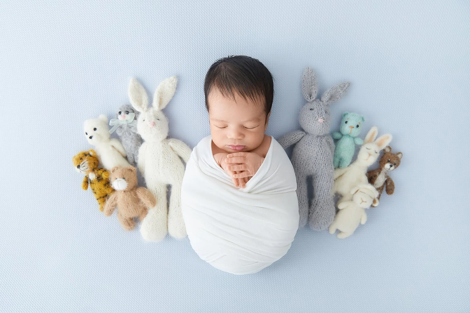 Swaddled baby surrounded by stuffed animal bunnies in pastel colors, on light blue background.