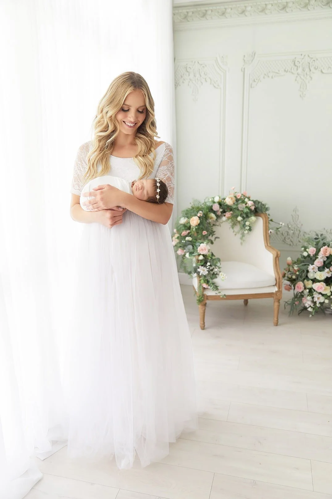 A woman in a white dress holding a newborn baby in a floral headband, standing in a bright room with floral decorations and a vintage chair.