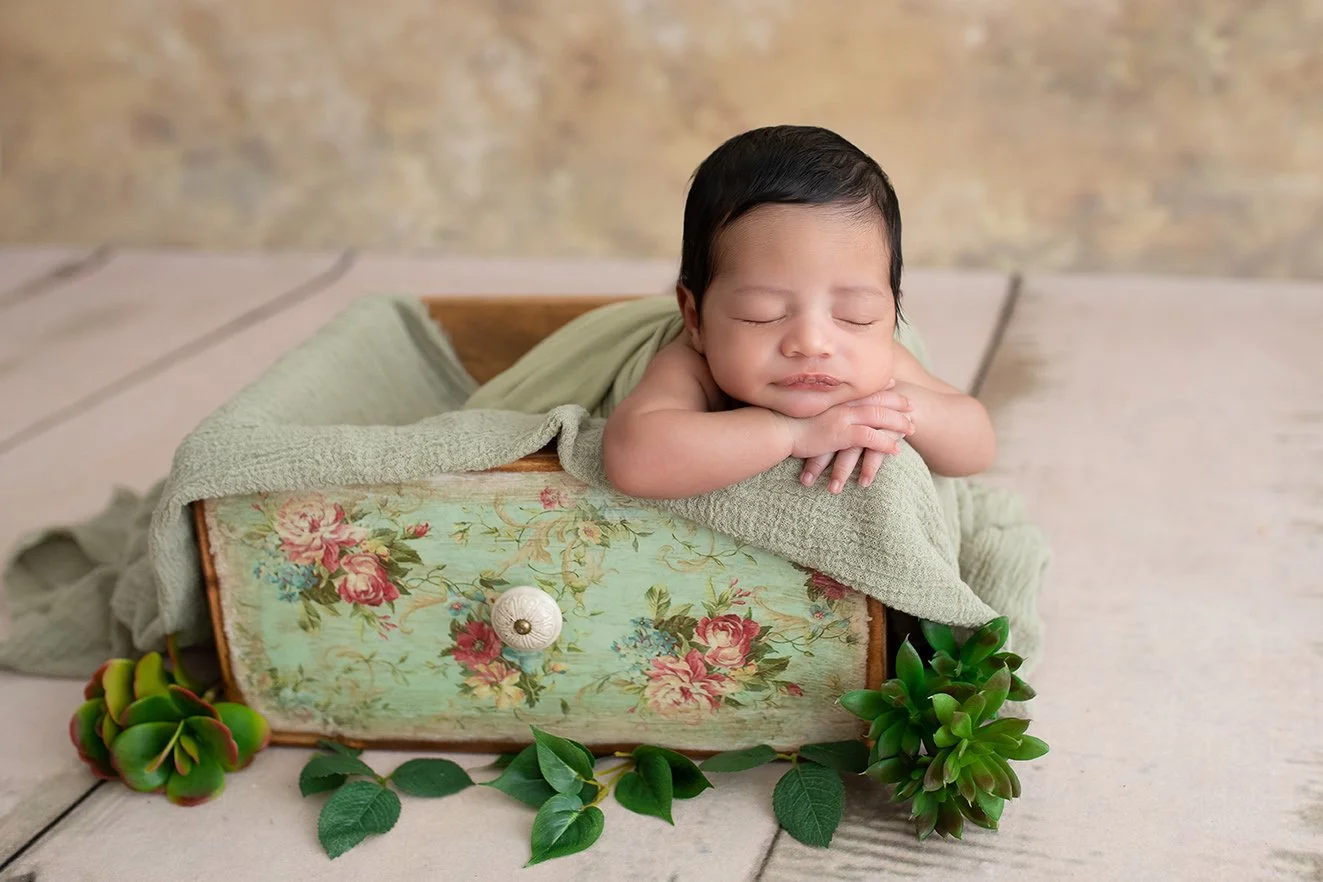 A sleeping baby with dark hair resting its head on its folded arms, lying in a wooden drawer decorated with floral fabric, with greenery and succulents around it.