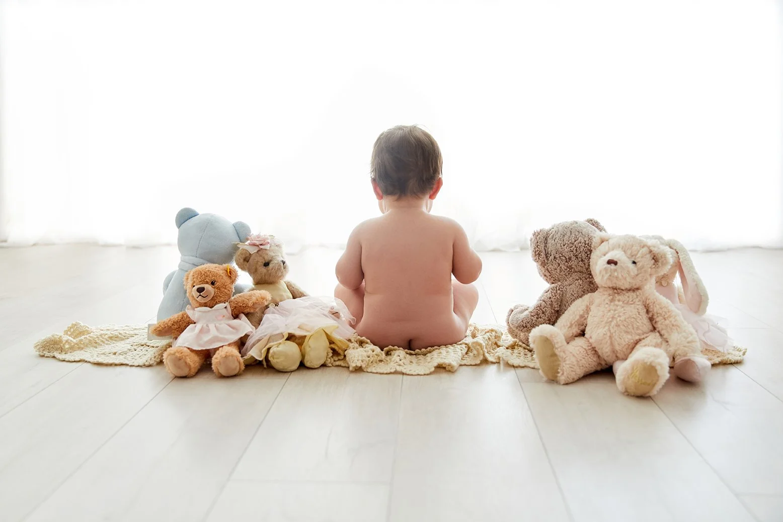 A baby sitting on a blanket, surrounded by stuffed animals including bears and a bunny, facing a bright window.