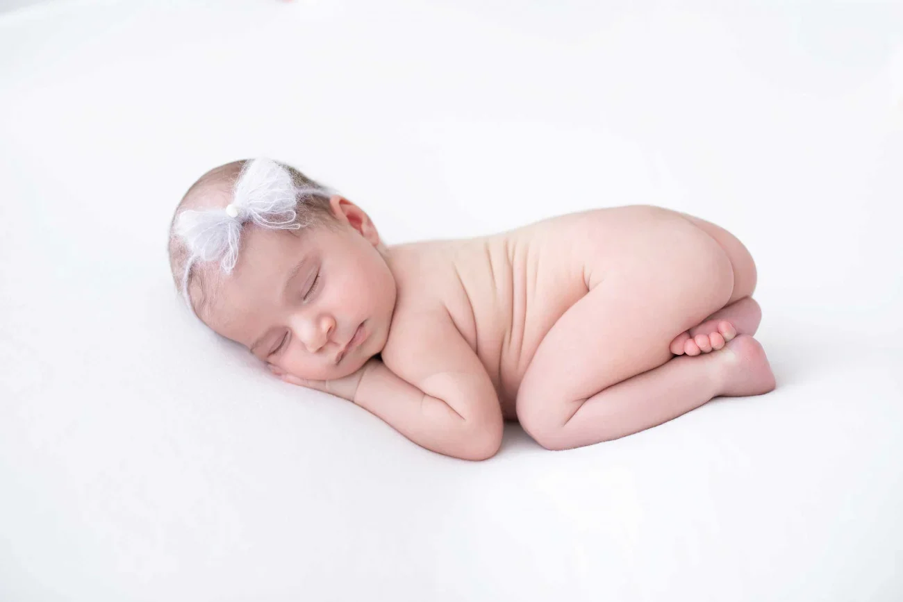 A sleeping newborn baby lying on its side on a white surface, wearing a white headband with a bow.