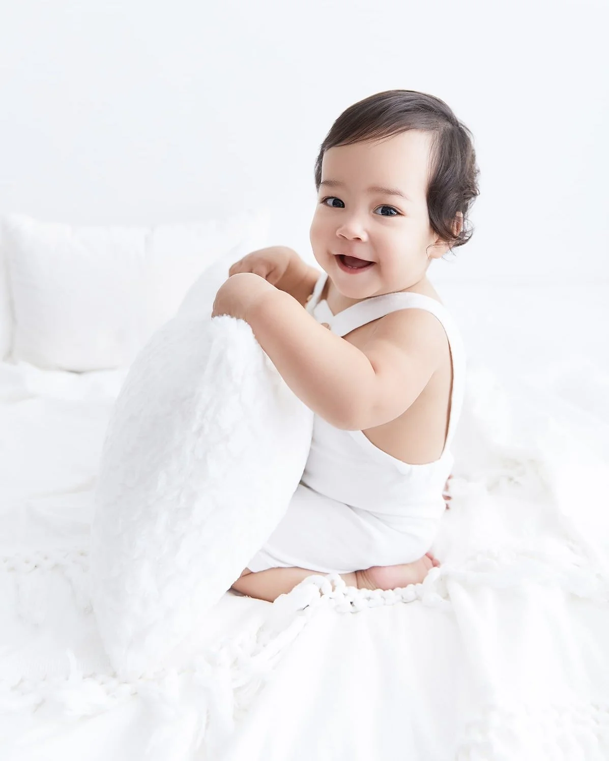Smiling baby with dark hair sitting on white bed, holding a white pillow in a bright room.