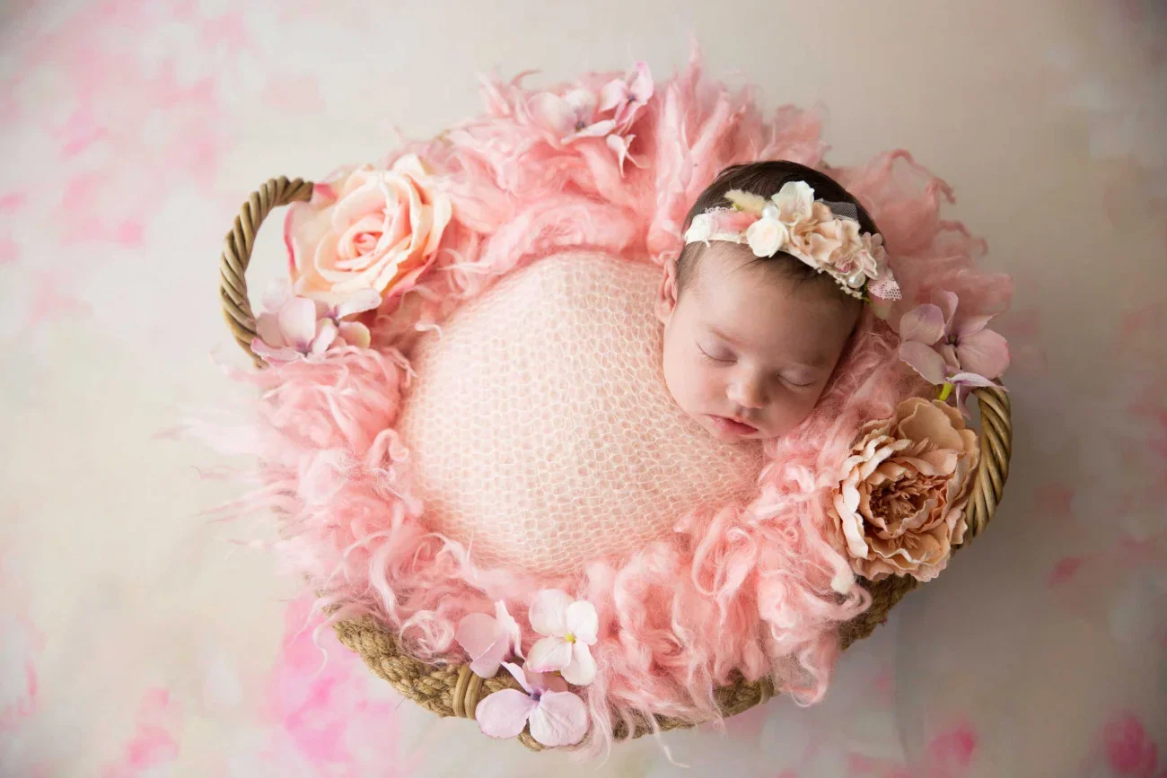 A sleeping newborn baby with a floral headband, wrapped in a pink blanket inside a woven basket decorated with pink and peach flowers and pink fluffy material.