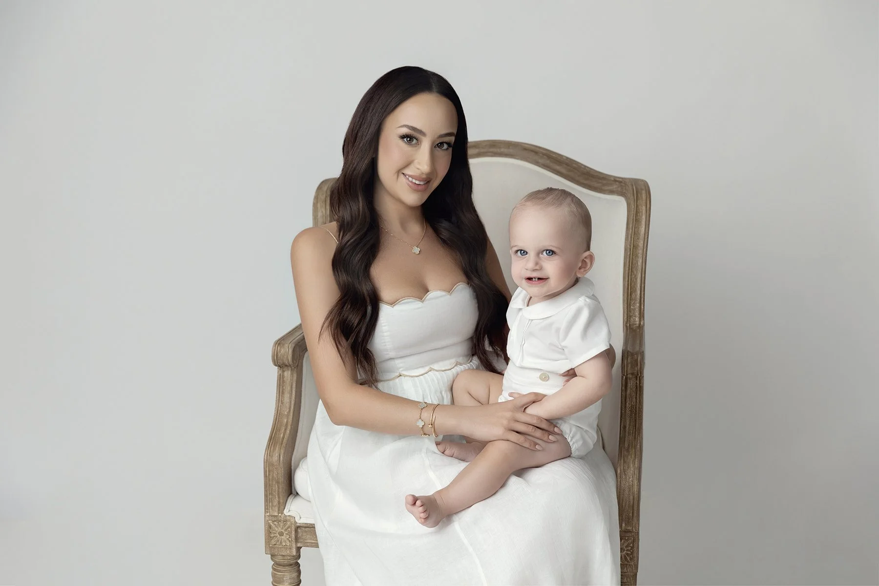 A woman with long dark hair sitting on an ornate wooden chair, holding a young toddler with light hair and blue eyes, both dressed in white against a plain light-colored background.
