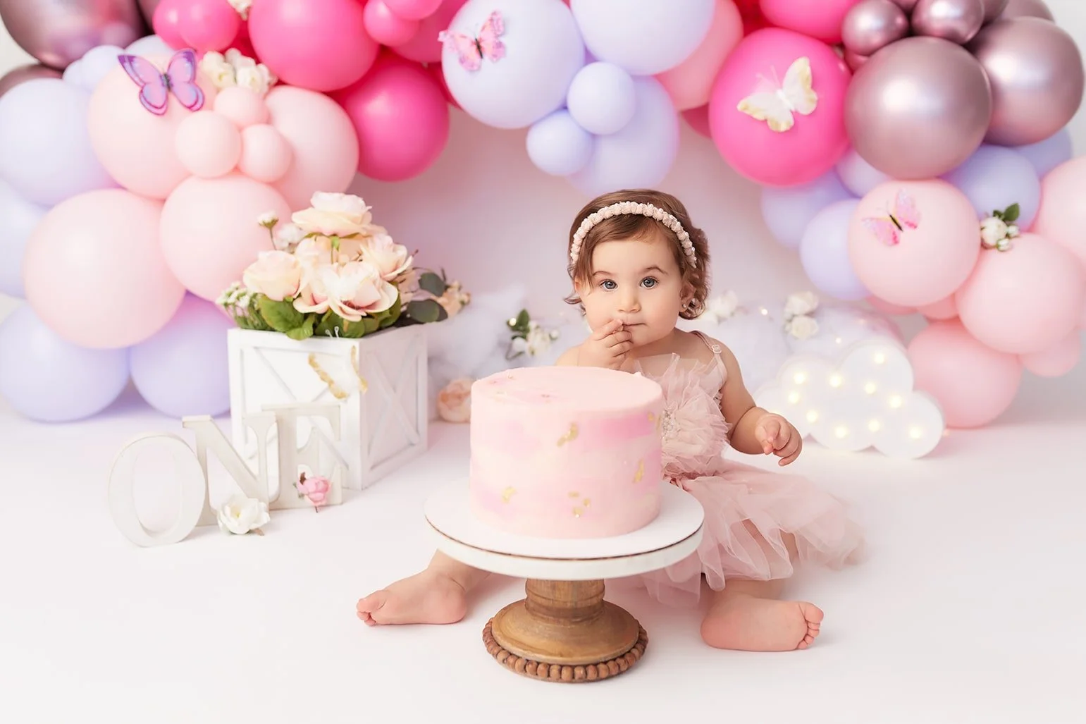 A toddler girl sitting on the floor in front of a pink birthday cake, with pink, white, and metallic balloons in the background, decorated with butterfly and flower embellishments.