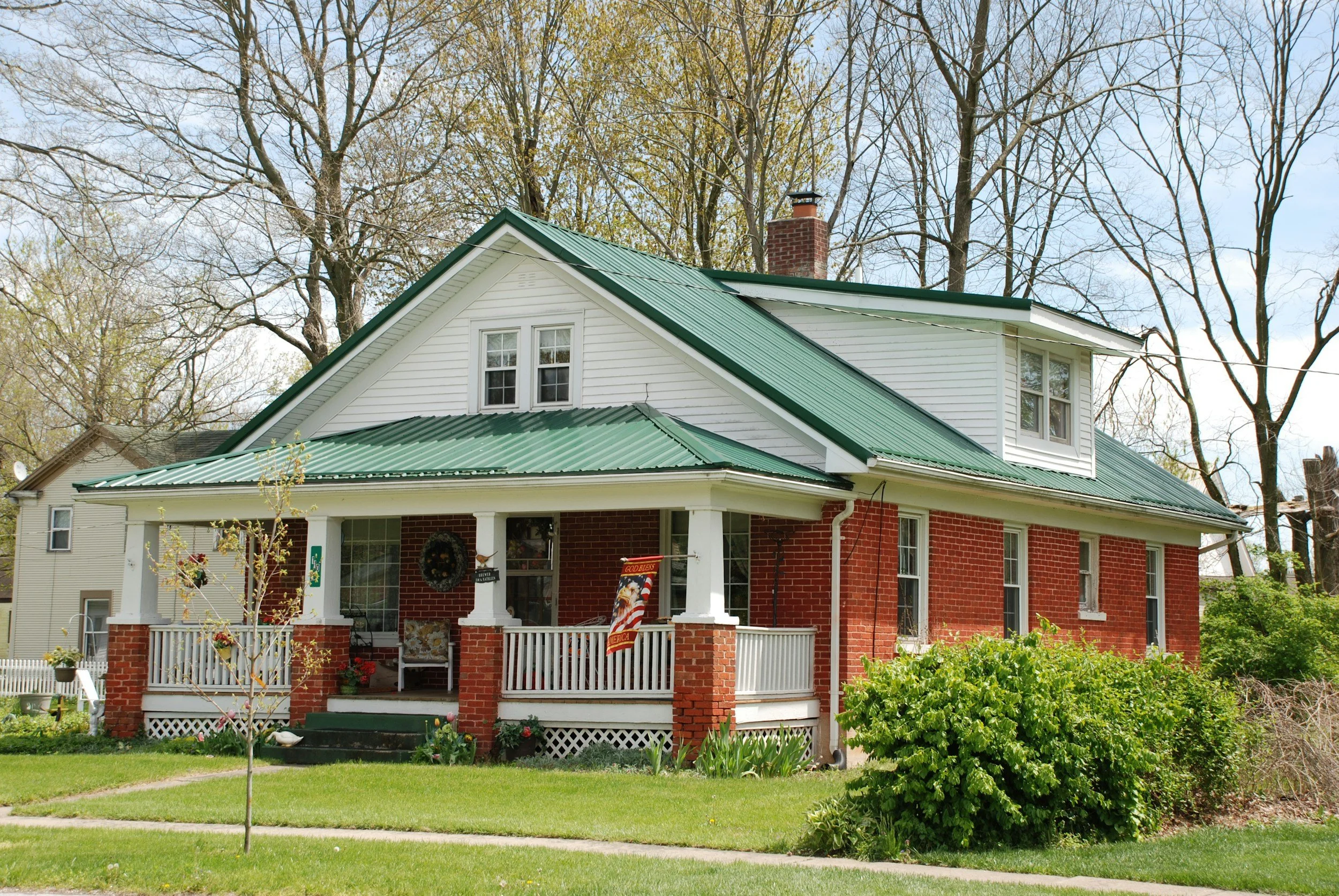 A house with red brick and white siding, green metal roof, front porch with white railing, surrounded by green lawn and trees.