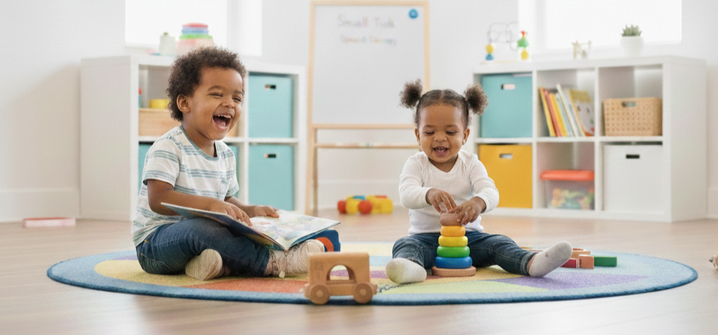 Two young children, a boy and a girl, sitting on a colorful round rug in a playroom, laughing and playing with toys. The boy holds a book, and the girl stacks colorful rings. Background includes shelves with books, toys, and small decor, with a whiteboard and windows.