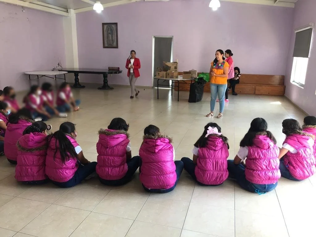 Niños y niñas sentados en círculo en un aula, con una mujer de pie frente a ellos y otras dos personas en la parte trasera, en un ambiente con paredes rosadas y ventanas con cortinas.