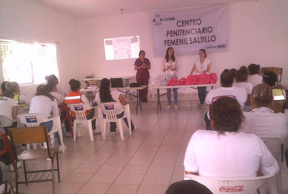 Reunión en un centro penitenciario femenino en Saltillo, donde varias mujeres y algunas en uniforme de trabajo están asistiendo, con tres mujeres de pie frente a una mesa, una de ellas hablando, y una pantalla proyectando una presentación, en un salón con paredes blancas y ventanas.