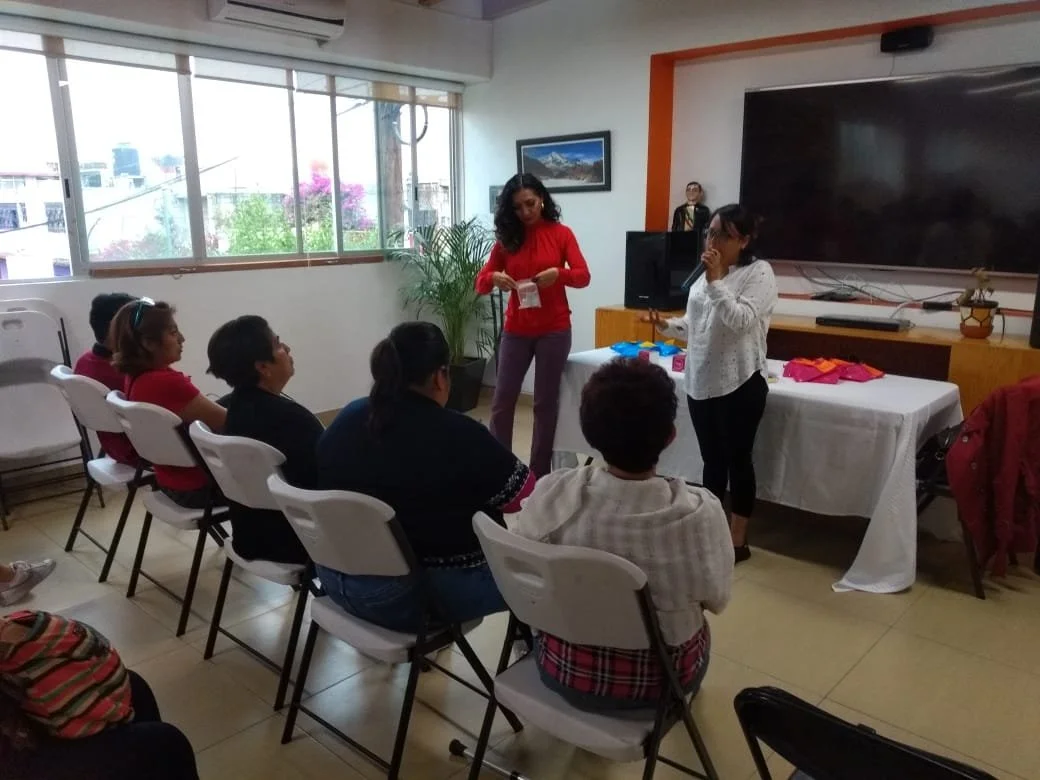 Grupo de personas participando en una reunión o taller en interiores, con dos mujeres de pie en frente, una entregando folletos y la otra hablando, mientras que varias personas sentadas observan.