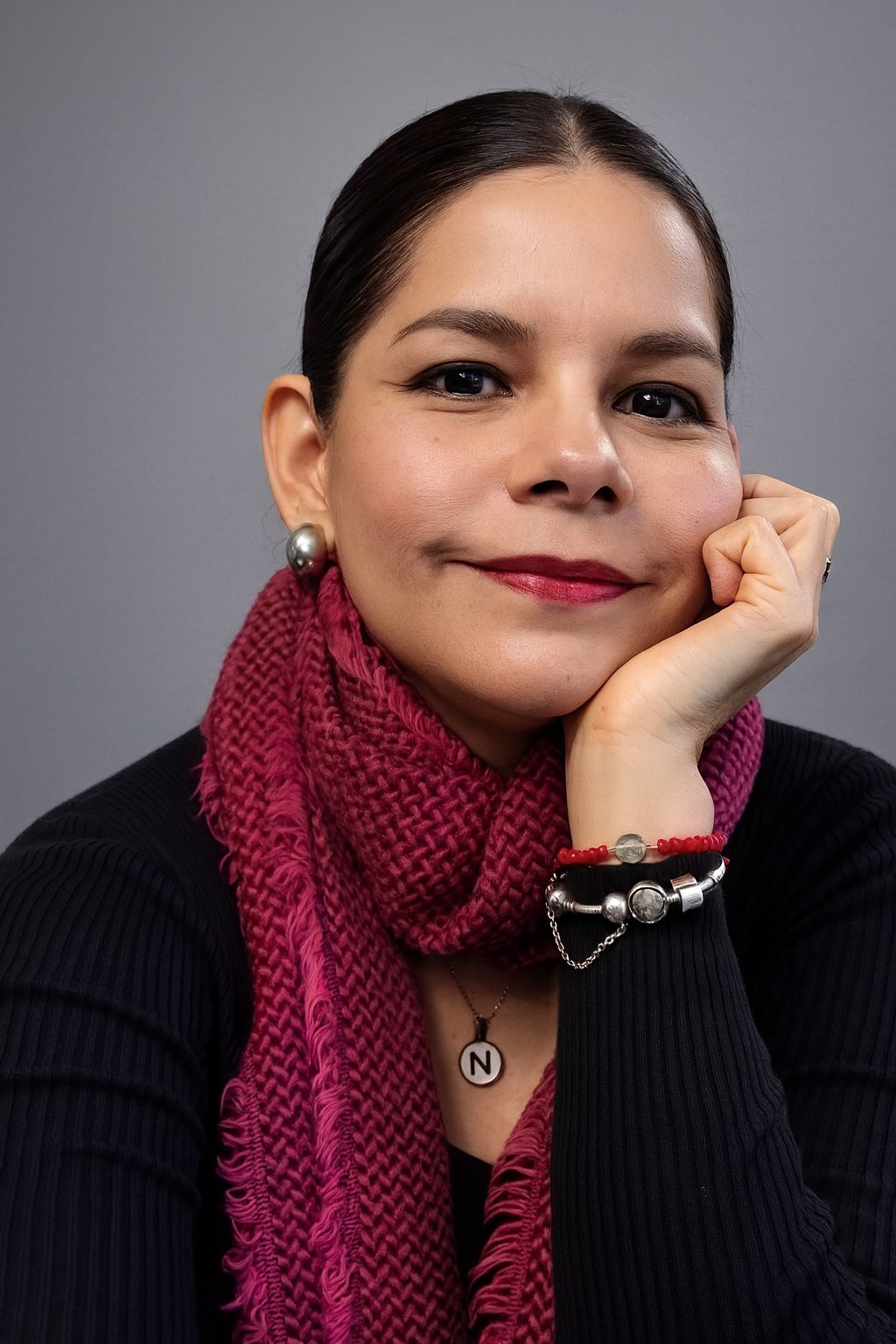 Retrato de una mujer con cabello oscuro peinado hacia atrás, vestida con un suéter negro, bufanda rosa, usa pendientes grandes de perla, un collar con una letra N, y pulseras de bolitas de colores, sonriendo ligeramente en un fondo gris.
