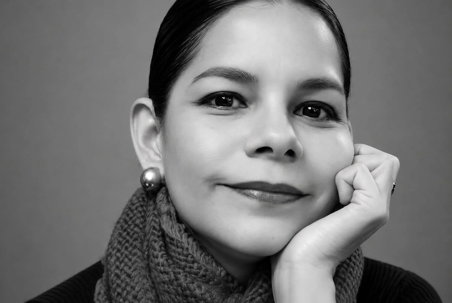 Retrato de una mujer con cabello oscuro peinado hacia atrás, vestida con un suéter negro, bufanda rosa, usa pendientes grandes de perla, un collar con una letra N, y pulseras de bolitas de colores, sonriendo ligeramente en un fondo gris.